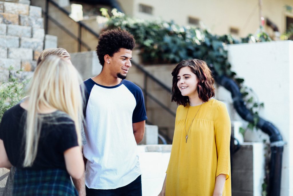 In front of a stone staircase a white woman with short dark hair wearing a yellow dress is talking to a black man with natural hair and a goatee wearing a white t-shirt with blue sleeves. A white woman with long blond hair and a plaid skirt stands in the foreground facing them