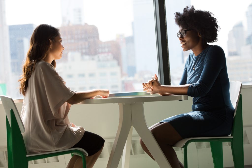 In front of a window looking out over a city scape, A white woman with long brown hair wearing a large pink t-shirt is sitting at a table across from a smiling black woman with natural hair wearing glasses and a navy blue t-shirt and gold necklace