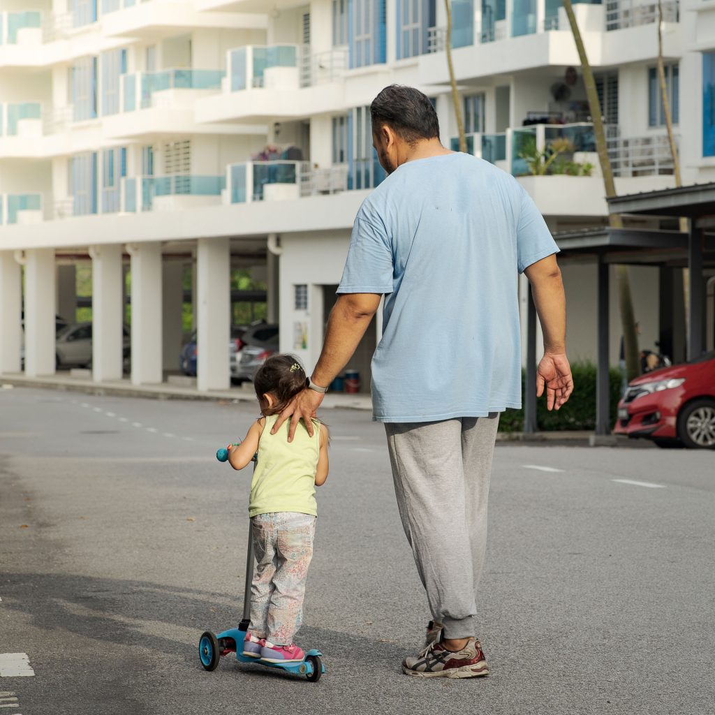 In the parking lot of an apartment complex, a Hispanic man in a blue shirt and grey sweat pants is assisting a small Hispanic girl wearing multicolored pants and a yellow tank to ride a blue scooter.