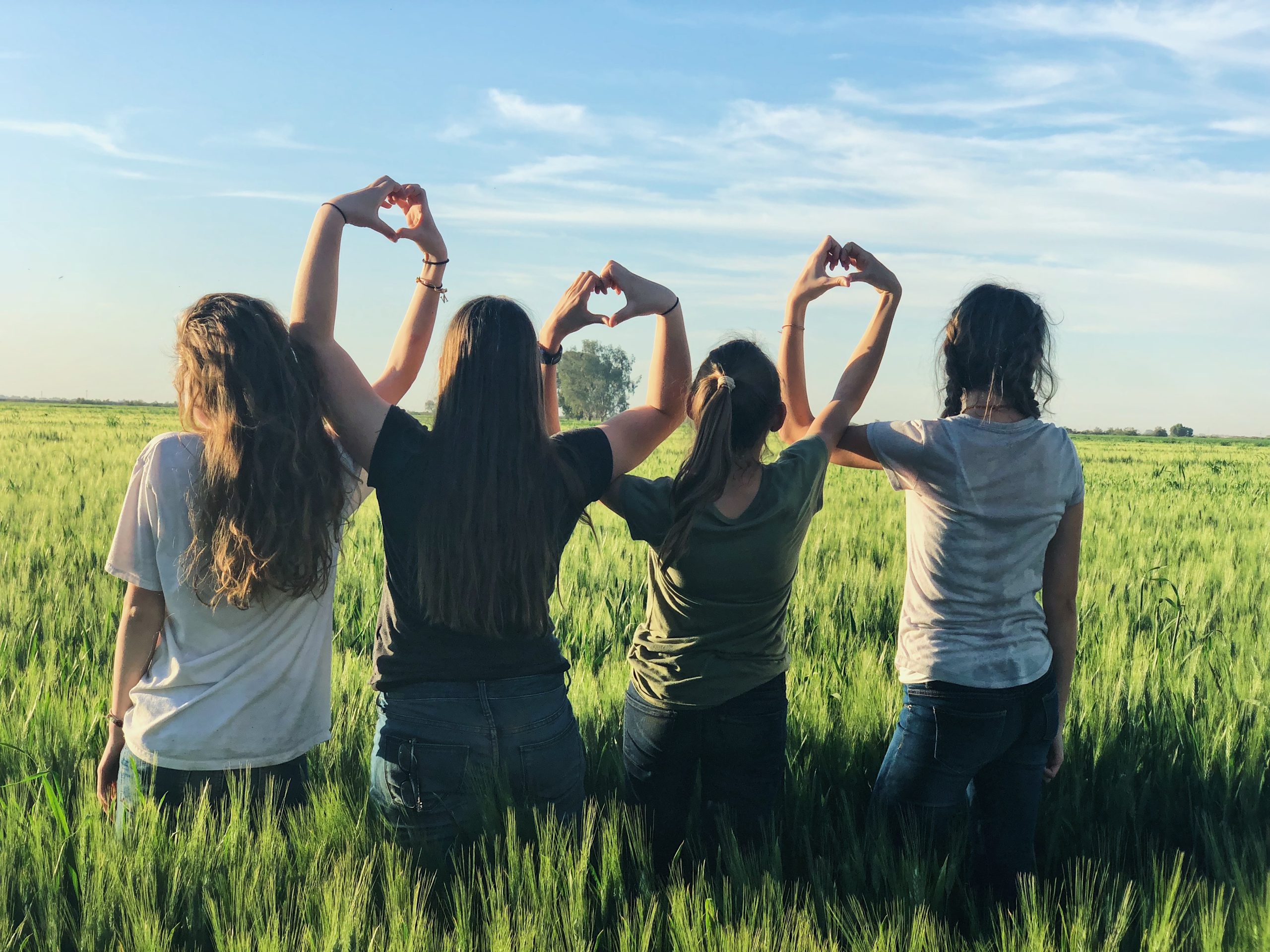Four young white women stand in a field of tall grass facing away from the viewer. Their arms are over their heads linking their hands in the Heart symbol