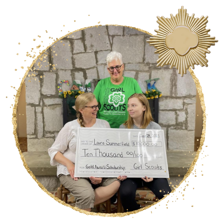 In front of a stone fireplace a white woman with white hair and glasses wearing a Green GIrl Scout shirt stands behind a seated white woman in a white top with a blond braid and a young white girl with strawberry blond shoulder length hair. The woman and the girl are holding a large printed check for 10 thousand dollars Gold Award Scholarship. A gold trefoil over a sunburst pattern is in the top right corner.