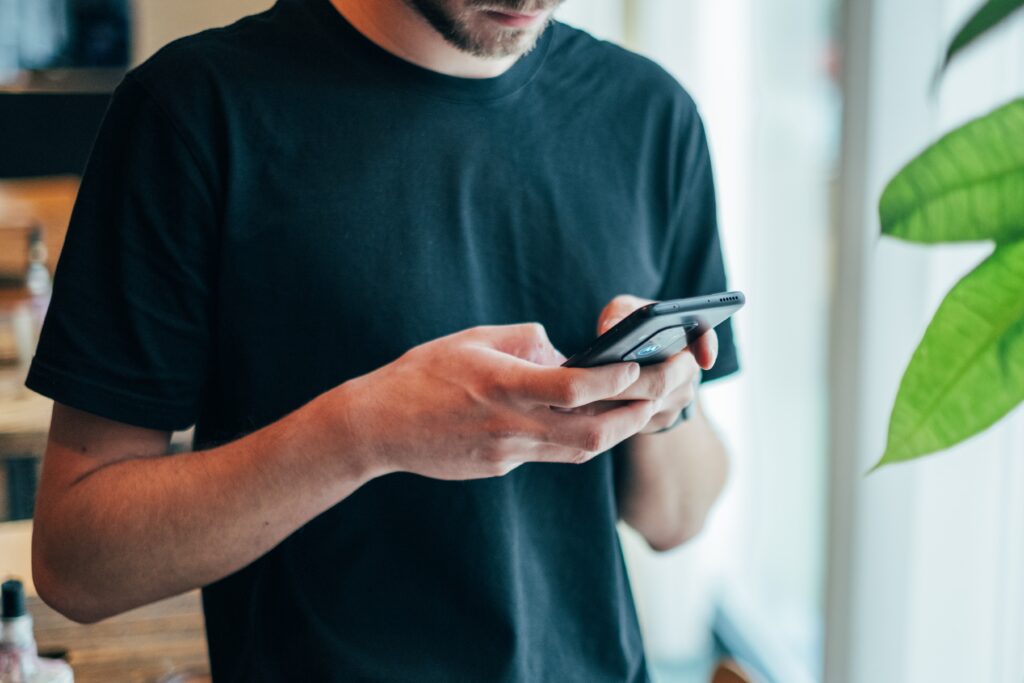 A young white man with a beard wearing a black t-shirt is looking down at a cell phone held in both hands.