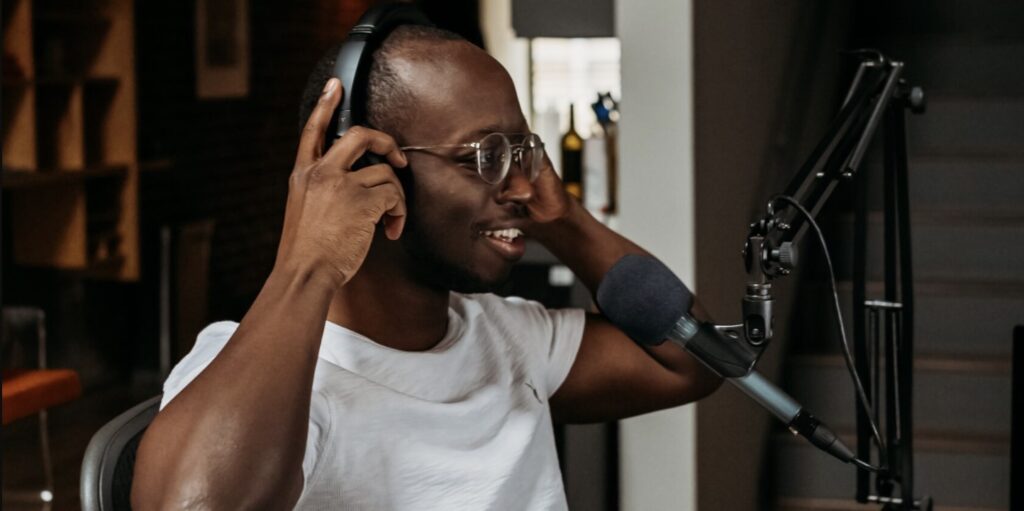 A black man in a white teeshirt and glasses is adjusting headphones over his head and is sitting in front of a microphone