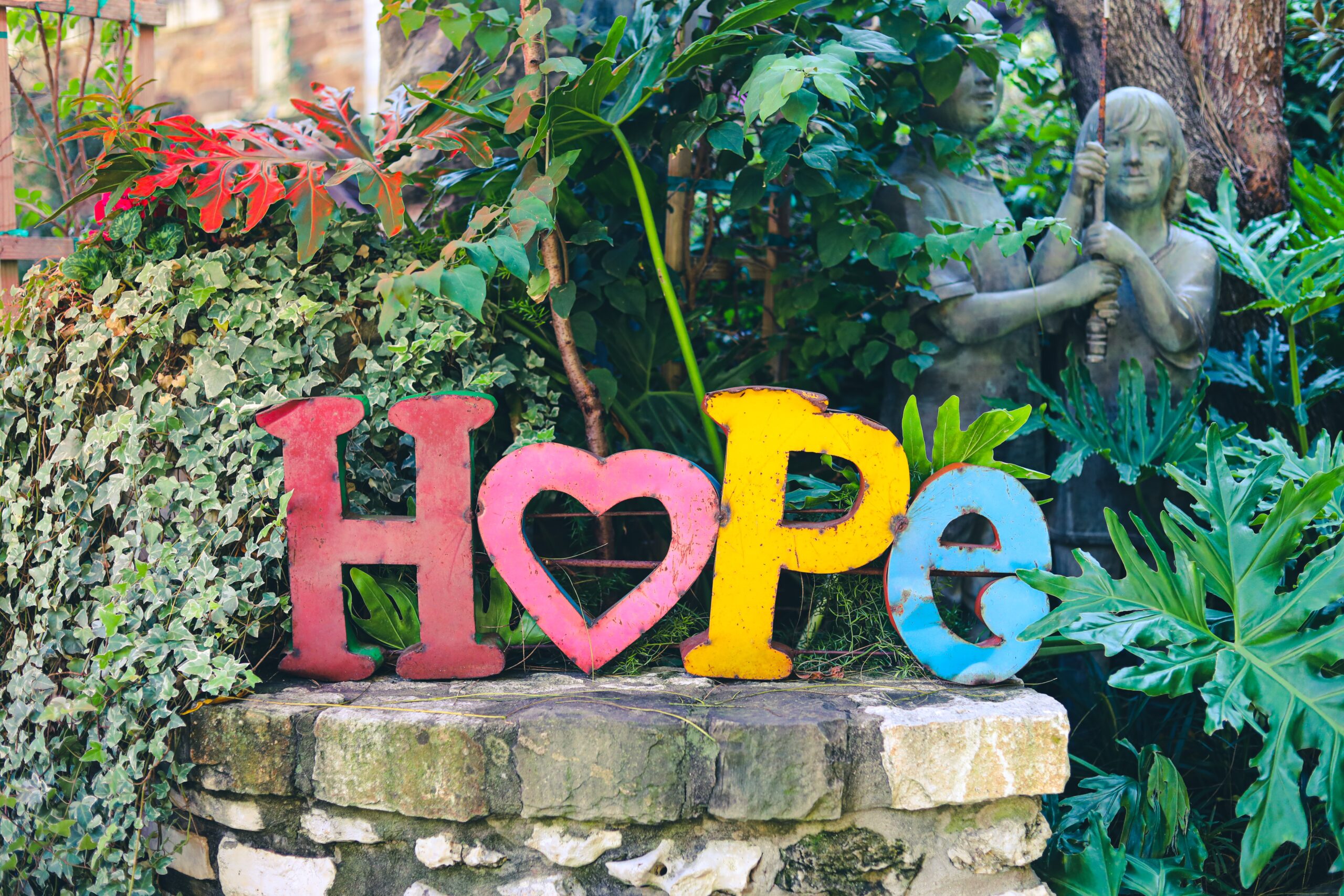 Against an ivy-coated wall, the word hope is spelled out in weathered metal letters. The O is represented by a heart. There is a stone statute of children in the background.