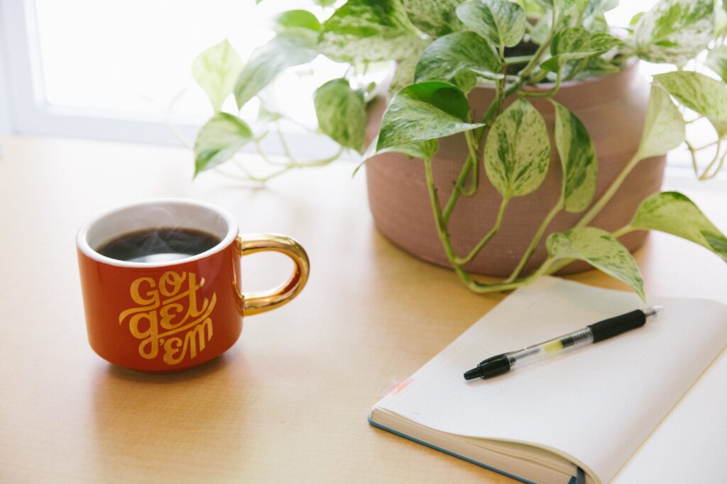 A desk in front of a sunny window. A houseplant sits in a light red pot. In front and to the left is a red steaming cup of coffee with the words Go Get 'em written in gold. In the foreground an open notebook has a pen on top of it.