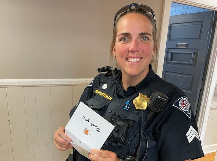 A white female police officer, with brown hair pulled back in a pony tail with sunglasses on her head. She is holding a Thank you note and smiling.