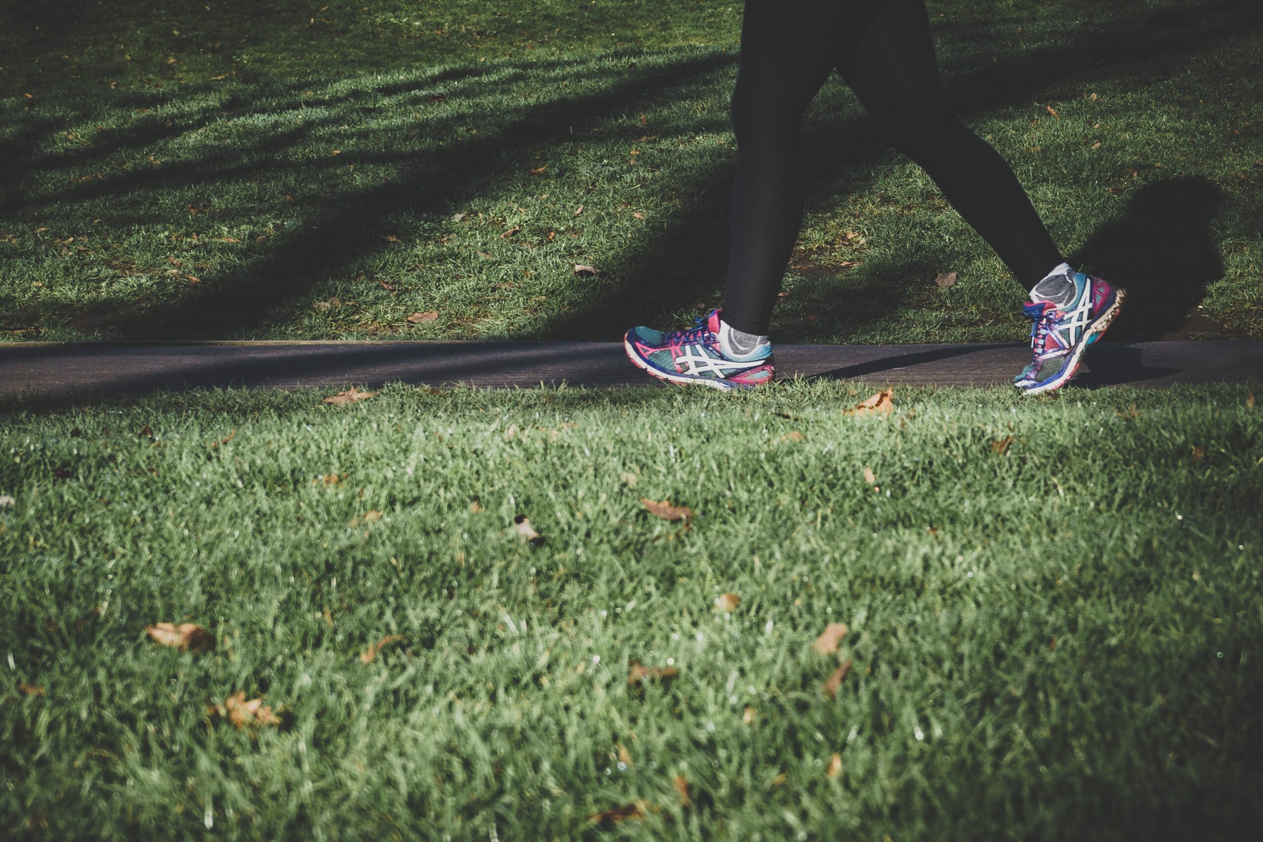 A persons legs from the hips down wearing leggins and sneakers walks on a paved path between patches of grass dotted with leaves.