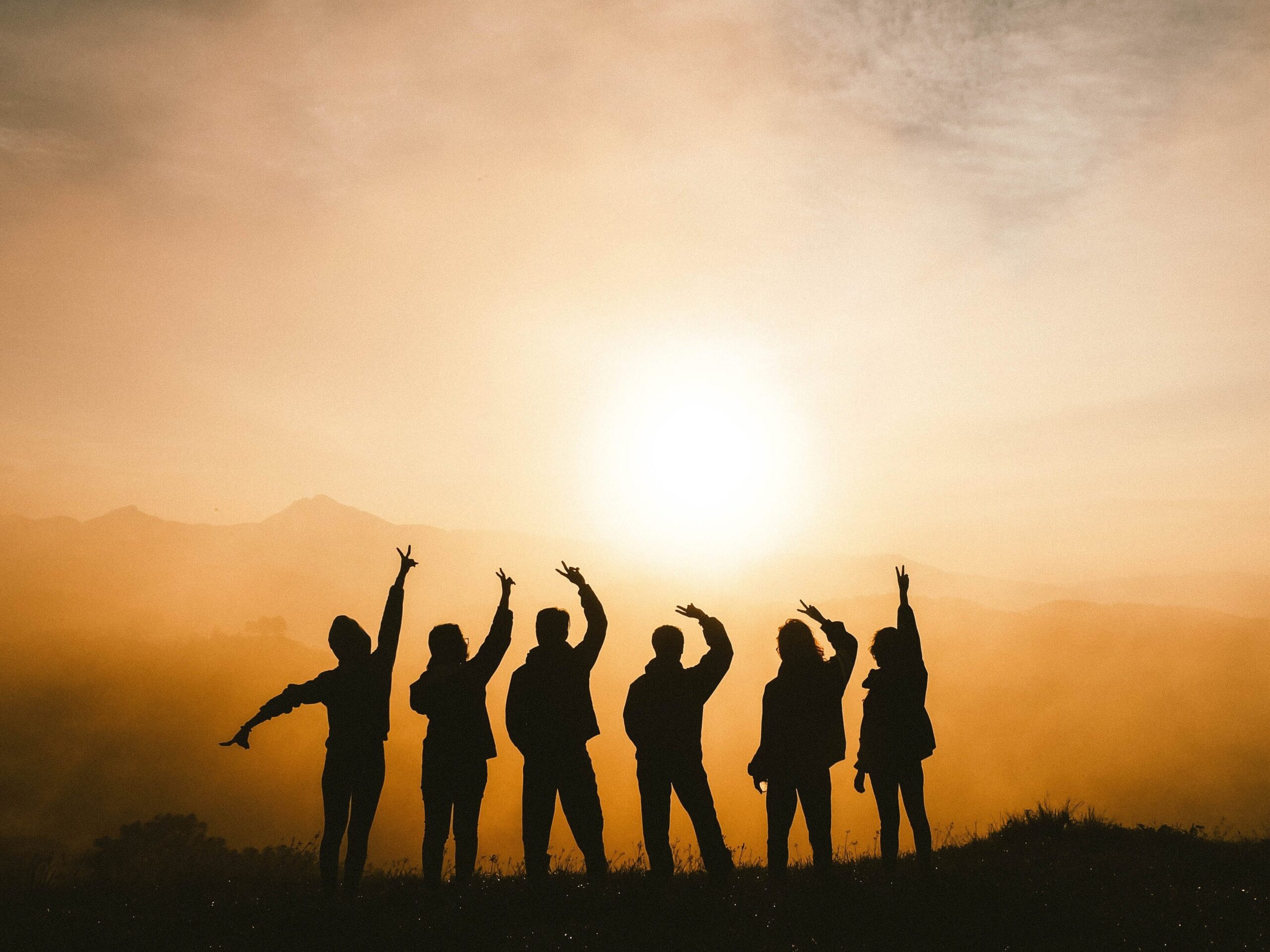 The sun is setting behind mountains letting off an orange light. 6 people stand in silhouette with one arm extended above their heads displaying the peace sign.