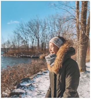 A woman in a brown fur hooded parka and hat smiles while looking at a lake, there is snow and winter bare trees in the background with a bright blue sky.