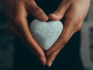 A person's hands are shown close up holding a heart-shaped stone with the word hope writtern on it.