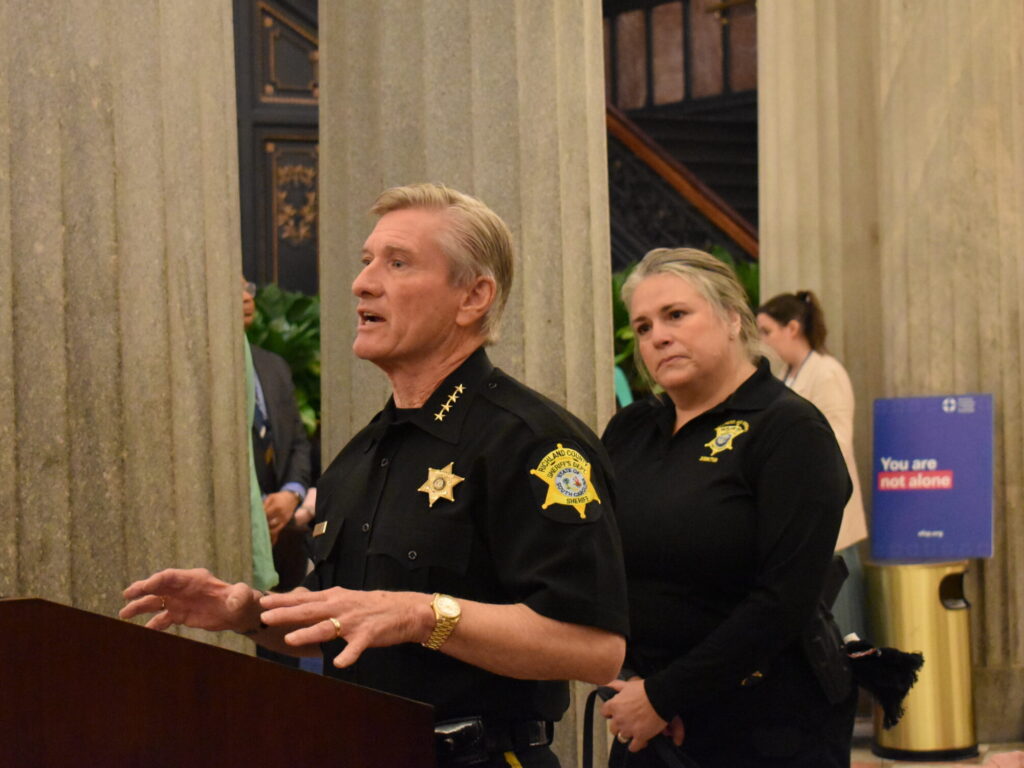 Sheriff Lott of the Richland County Sheriff's Department stands before a podium speaking to an unseen crowd. An officer stands behind him observing.