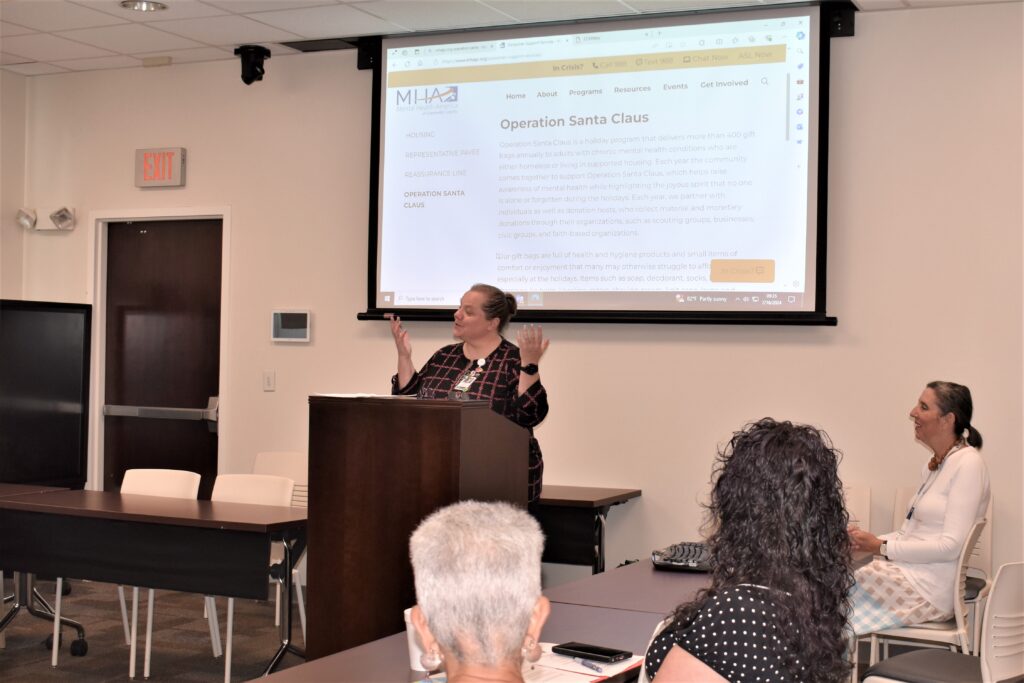 A picture of a white woman in a black and red plaid shirt speaks at a podium in front of a drop down screen that says Operation Santa Claus.