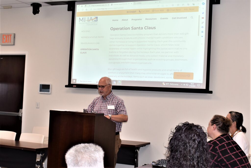 A photo of a white man in a red plaid shirt speaking from a podium in front of a drop down screen with the words Operation Santa Claus.