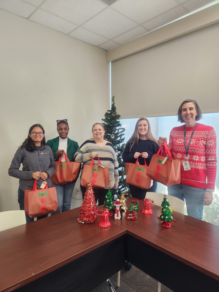 Five Smiling women holding Reindeer gift bags stand in front of a Christmas Tree