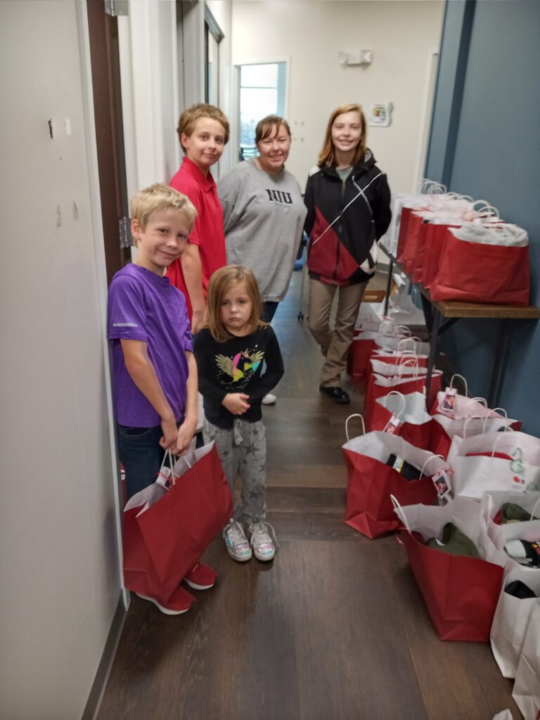 A group of children stand in a hallway surrounded by gift bags that they have helped pack.