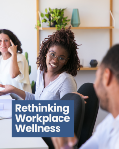 A smiling young black woman with twists in her hair and glasses is talking to her coworkers in a brightly lit office.